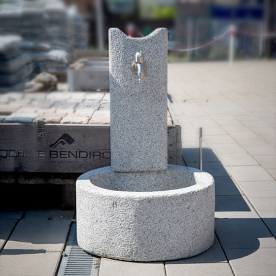 Granitbrunnen aus hellgrauen Stein mit einem edelstahlfarbenen Wasserhahn verkauft für den Garten von Löseke´s Steingarten.